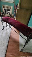 Long view of bench showing black wrought iron frame with ornate scrollwork, and long red upholstered seat showing dust marks and wear.