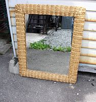 Full view of the large rectangular wall mirror with woven thick woven straw surround, standing upright outdoors against a white wall and concrete ground.