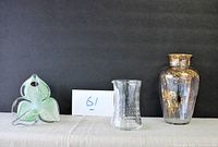 Three pieces displayed side by side: green and white floral Murano glass sculpture with spiral stem, clear textured glass vase, and Murano hand painted gold flower vase
