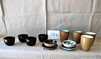 Photo of all twelve assorted ceramics including black tea cups, textured brown cups with colored interiors, and small blue/white bowls with saucers.