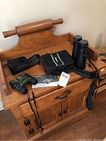 Two sets of binoculars placed on a wooden desk. The Celestron Outland X 10x50 binoculars with black carrying case, folded instruction manual, and a tag. The Barska binoculars in black and green colors are also visible.