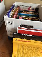 Box of assorted hardcover and softcover illustrated books showing various colorful titles on comics, cult cinema, horror, and men's magazines.