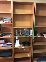 Three tall pressboard bookcases with adjustable shelves shown partially filled with various items. Middle bookcase has electronic devices, bells, and miscellaneous items temporarily placed on shelves.
