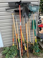 Multiple long-handled garden tools leaning against siding. Includes rakes, shovel, broom, cultivator and more.