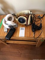 Photo shows four items on wooden surface: white Beats by Dr. Dre headphones folded, beige and black Sennheiser HD598 wired headphones with cable, Sony USB recorder, and Logitech webcam with clip.