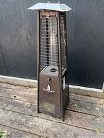 Full view of the standing outdoor propane heater with glass chimney, metal cage, and top shield on wood deck with black wall background.