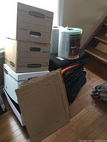 Photo of five cardboard bankers boxes stacked near window, with brown folding box pieces in front, and underneath several folded moving blankets with two rolls of protective self-stick wrap on top.