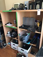 Shelf with various office and electronic accessories including power cables, bookends, headphones, and multiple small devices in plastic bins and boxes.