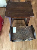 Top view of dark wooden stool seat showing saddle shape and recessed square accents, with visible scratches and wear. Wooden wine rack partially visible beside stool.
