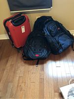 Photo of three bags on a wooden floor; red hard case suitcase, black Swiss Army backpack, and blue Samsonite backpack.