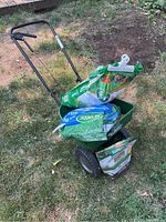 Photo of green Scotts Turf Builder Edgeguard Mini spreader with handle and wheels, holding bags of grass seed and fertilizer on the grass.