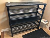 Photo of one black metal shoe rack with three tiers of parallel metal rods and a flat rectangular top against a beige wall on tiled floor.