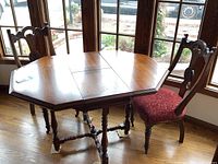 Octagonal wooden antique table with two matching chairs positioned near large windows on hardwood floor.