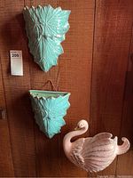 Ceramic collection on wood wall showing two turquoise leaf ceramic wall pockets and pink swan figurine