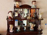 Full shelf view showing 11 cameo girl head vases arranged on a wooden mirrored shelving unit for scale and quantity reference.