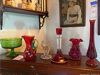 Collection of six hand blown glass pieces displayed on wood surface: green pedestal bowl, red pitcher, clear angel figurine, and three red hobnail vases.
