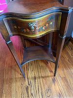 Full view of wood side table showing curved front drawer with floral hand painted design and drawer knob, lower shelf, and wood grain surface.
