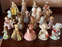 Photo showing the full assortment of 19 porcelain or ceramic figurines in pastel and gold tones arranged on a wooden surface.