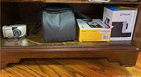 Kodak EasyShare M643 camera box, Polaroid OneStep 2 Viewfinder & Color Camera box, black camera soft case, and one Olympus camera on wooden shelf.
