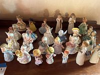 Wide view showing 25 assorted angel figurines on a wooden surface against a beige wall. Various colors and poses visible including holding objects and some with halos.