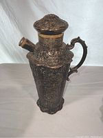 Two views of an antique silver plated cocktail shaker showing ornate embossed detail, curved handle, hinged lid and spout, with some tarnish.