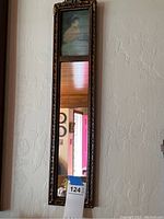 Full vertical view of the narrow rectangular mirror with ornate frame hanging on a textured wall, showing the reflection below the illustration.