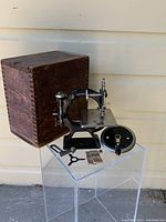 Wide view of machine, wooden box, hand crank wheel, bracket, needle packet and clamp on clear stand