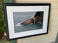 Framed large format photograph showing Newfoundland coastline with cliff, road, building, and ocean.