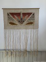 Full view of the vintage textile wall hanging showing the woven wool geometric design and hanging yarn fringes against a white wall.