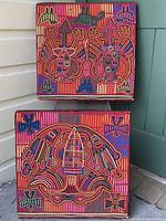 Photo showing two mounted square mola textiles with intricate multi-colored patterns and animal designs, photographed against exterior wall corner