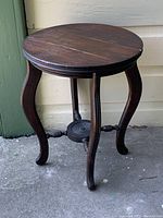 Wooden round plant stand on concrete floor next to green siding, showing top, legs, and stretcher base.