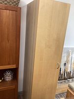 Front view of tall single door cabinet with light wood finish, closed door showing silver curved handle.
