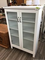 Front view showing white cabinet with frosted glass doors and metal handles, displaying three shelves inside.