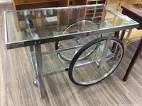 Front angle view showing large bicycle-style wheels on one side and chrome frame with glass shelves