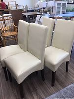 Four cream faux leather dining chairs arranged in two rows on dark wood floor in showroom setting.