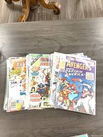 Photo showing a stack of Avengers comic books on a wooden table, covers featuring Captain America and other Avengers characters.