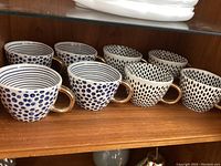 Two sets of four ceramic cups arranged in a row on a wooden shelf. The front set has blue polka dots, and the back set has blue concentric circles. Each cup has a gilt handle.
