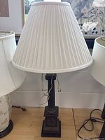 Full lamp showing white pleated fabric shade and decorative dark metal base, with cord visible at base.