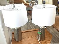 Photo showing side by side pair of table lamps with hammered metal bases and white fabric shades sitting on a wood surface.