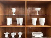Overall display of milk glass including goblets on top shelf, tumblers on middle shelves, cups and oval plates on lower shelf.