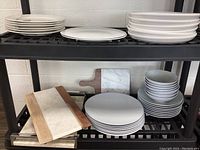 White Casa Blanca plates and other dinnerware stacked on plastic shelving, with wooden and marble charcuterie boards visible.
