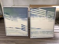 Pair of abstract canvas prints in white frames resting on floor, showing overall condition and color palette.