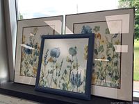 Three framed floral prints arranged together near a window, two larger prints with cream mats and light wood frames and one smaller framed in blue wood.
