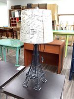 Full view of black metal Eiffel Tower style table lamp with fabric shade featuring script text design.
