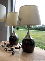 Pair of matching table lamps with dark brown ceramic bases and white fabric shades shown on a wooden surface near window.