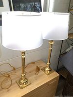 Pair of matching brass table lamps with white drum shades on wood surface showing full height and overall appearance.