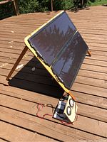 Solar panels mounted on angled wooden frame with support legs on a wooden deck. Connected to wiring and devices on the ground.