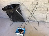Two foldable metal trash bag stands with one fitted with a black trash bag and a partial box of trash bags beside them.