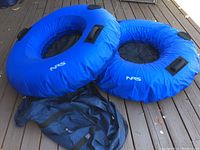 Two blue NRS Wild River float tubes shown partially inflated and fully inflated, lying on wooden surface with the included black nylon duffel bag beside them.