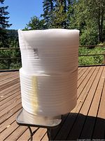 Two large white rolls of foam packing material shown upright on a glass-top metal table outdoors on a wooden deck.